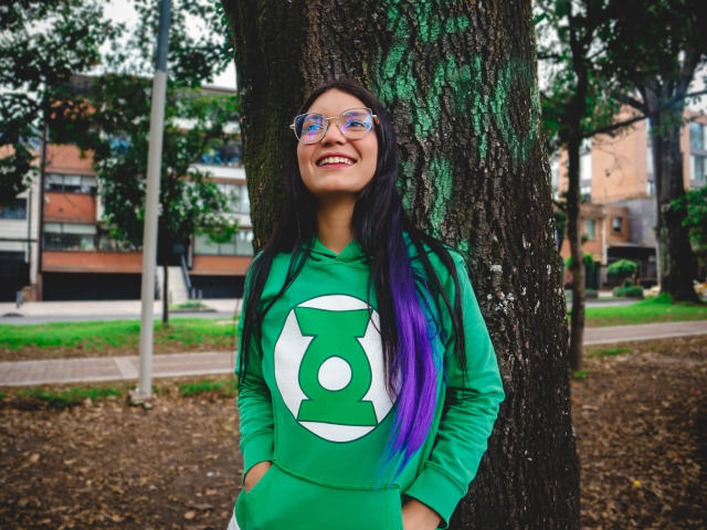AriaLeenn está em frente a uma árvore, vestindo um moletom verde com um círculo branco e um símbolo verde, cabelo roxo e preto, óculos azuis, sorrindo com as mãos nos bolsos..