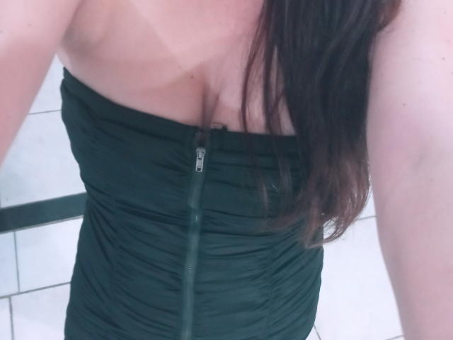 LadyNandas wearing a green, ruched, strapless dress with a visible zipper down the front, showing her boobs, with long brown hair falling on her right shoulder.
