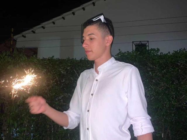 ThomasTwink69, con cabello negro corto, camisa blanca, sosteniendo una chispa encendida, con una ligera sonrisa, fondo oscuro, usando gafas de sol en la cabeza..