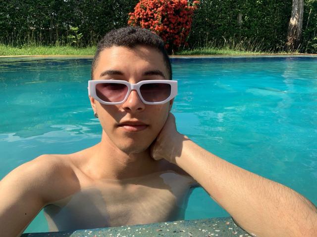 ThomasTwink69, avec des cheveux courts et bouclés, une peau bronzée, de grandes lunettes de soleil blanches et sans chemise, la tête reposant sur sa main, dans une piscine bleue, regardant l'appareil photo, avec un buisson rouge en arrière-plan..