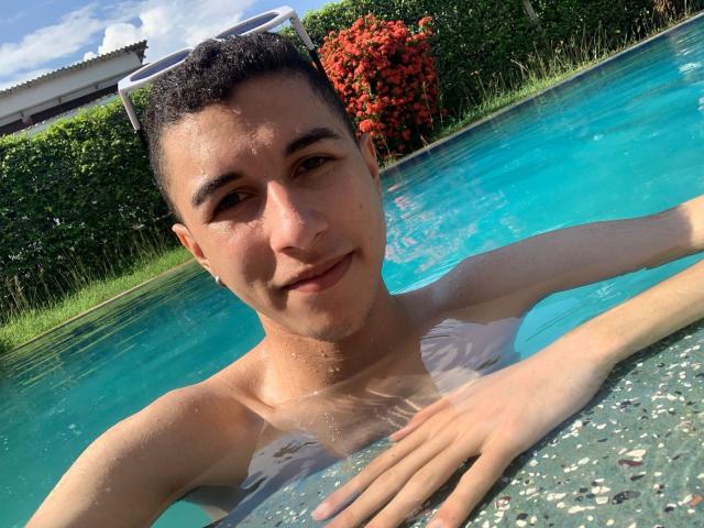 ThomasTwink69 im Pool, ohne Hemd, mit kurzen, dunklen Haaren, heller Haut und einem leichten Lächeln, die Hände ruhen am Rand, Wassertropfen auf seiner Brust, blauer Himmel darüber..