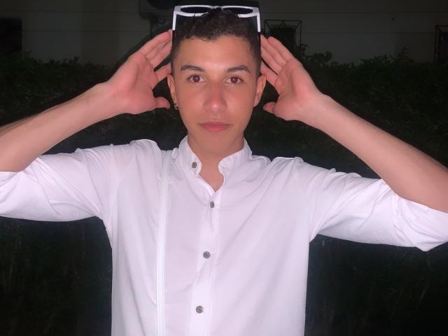 ThomasTwink69, vêtu d'une chemise blanche, porte des lunettes de soleil sur sa tête à deux mains, a les cheveux courts et foncés, et une expression neutre. Ses fesses ne sont pas visibles..