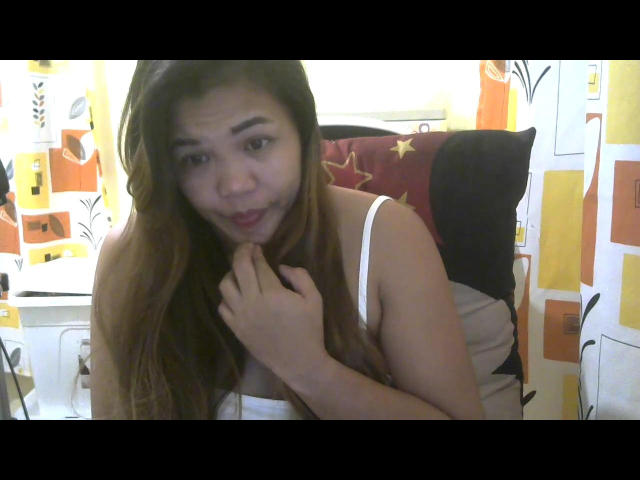 SweetJolieNikki with long brown hair, white tank top, and cute expression, biting finger, looking playful. Her hand touches her chin, showing off her tits subtly.