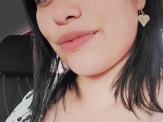 ElluzGabriela's face with black hair, pink lips, and a heart-shaped earring, showing a subtle smile. Her nose is small and she's wearing a white, black-patterned top.