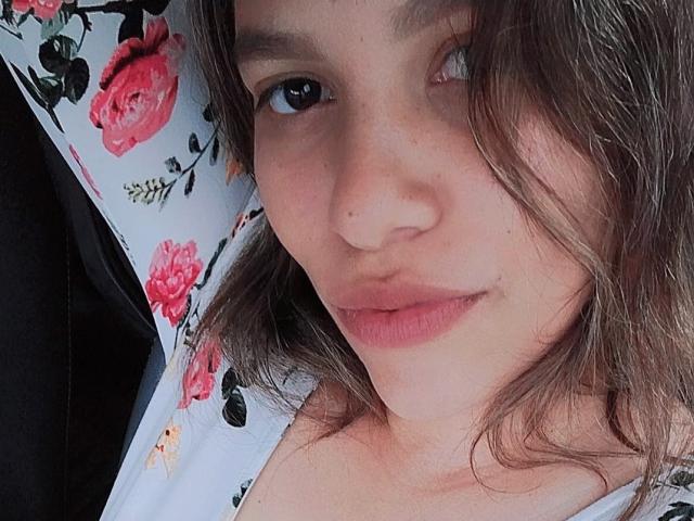 ElluzGabriela has light brown hair, a small nose, and pinkish lips. She wears a floral shirt with red roses. Her blue eyes look slightly to the side.
