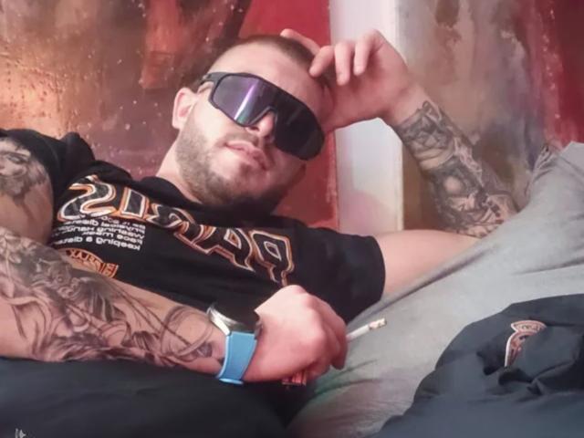 MiguelHot69, tattooed dude with dark sunglasses, black "21" shirt, blue watch, holding cigarette, lying back, smirking, right arm tattooed, left hand on head, gray pants, red wall.