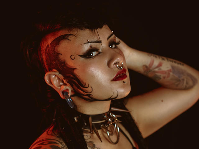 DafneLi with short black hair, red highlights, facial piercings, tattoos, black choker, blue ear gauges, hand in hair, red lipstick, dark background, moody lighting, serious expression.