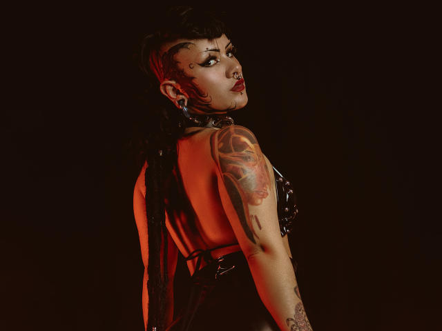 DafneLi with dark, thick dreadlocks, side-boob, nose ring, and tattoos on her right arm, wearing a black, shiny, backless top, looking over her shoulder. Red lighting highlights her.