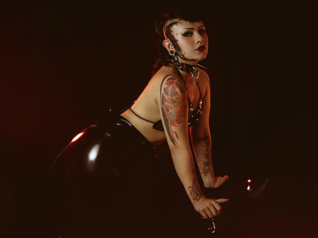 DafneLi in black latex pants and a leather top, on all fours, showing tattoos on her arms, with a nose ring and dark makeup, in a dimly lit setting.