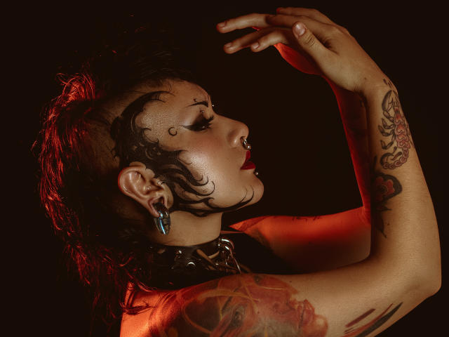 DafneLi with dark red hair, tattoos, black face paint, nose ring, earrings, and collar, raising arms, red light on skin, intense side profile, dark background.