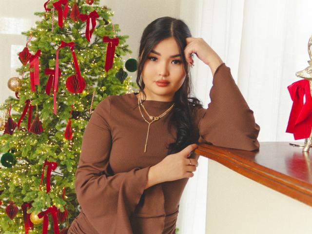 TeyaFaber with long black hair, brown long-sleeve dress, gold necklace, leaning on wooden table, next to decorated Christmas tree with red ribbons and green ornaments.