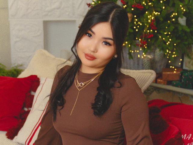 TeyaFaber with long black hair, brown long-sleeve top, gold necklace, red lipstick, and a cute smile, sitting on a red and white cushioned couch, looking at the camera.