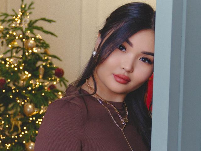 TeyaFaber with dark hair, red lipstick, brown top, pearl earrings, and necklace, peeks from doorway, Christmas tree with lights and ornaments in background.