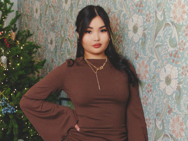 TeyaFaber with medium tits, brown dress, long brown hair, gold necklace, hand on hip, standing next to decorated Christmas tree.