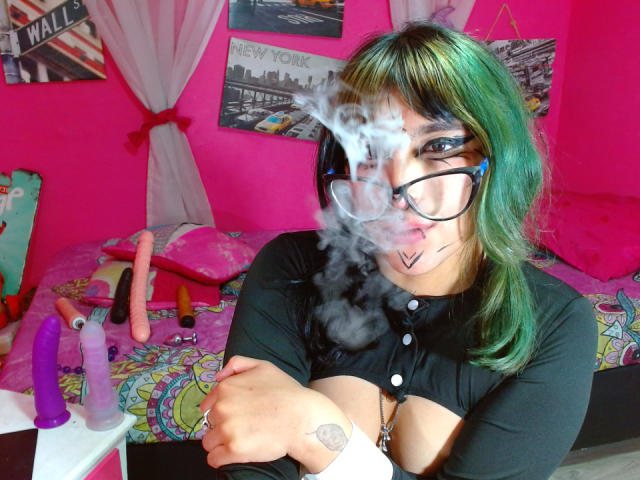 SerineHotAss with green and blue hair, black glasses, and a black top, has a cloud of cum on her face, with colorful bed and sex toys in the background.