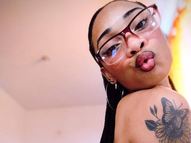 ElaHott pouts with red lips, glasses, and braids, showing a butterfly tattoo on her shoulder. Her brown skin and hoop earrings are visible. Her tongue is slightly out.
