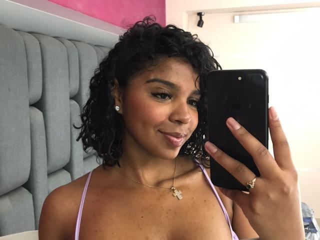 LisaMendez with curly black hair, brown skin, wearing a purple top, holding a black phone, taking a mirror selfie, showing off her small tits and a gold necklace.
