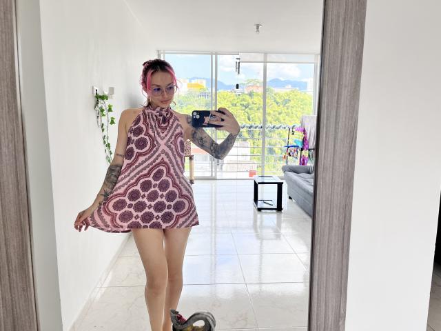 SarahRousen with pink hair, tattoos, wearing a pink, circular-pattern dress, standing in front of a mirror, holding a phone, showing her bare, cute, small ass and pussy.