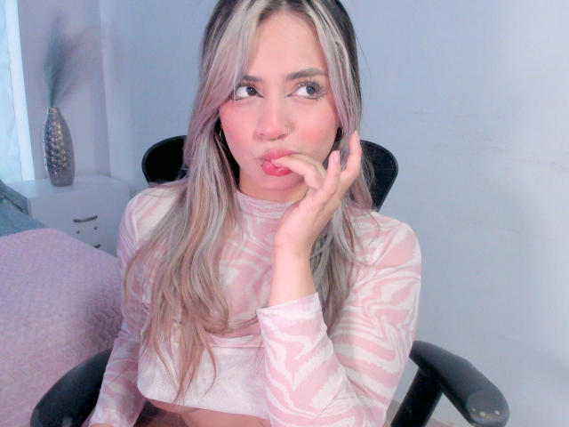 AlexaThoms, pouty lips, pink-striped long-sleeve top, blonde hair, nose ring, hand on cheek, pink lipstick, sitting in black chair, soft pink wall, subtle makeup, cute.