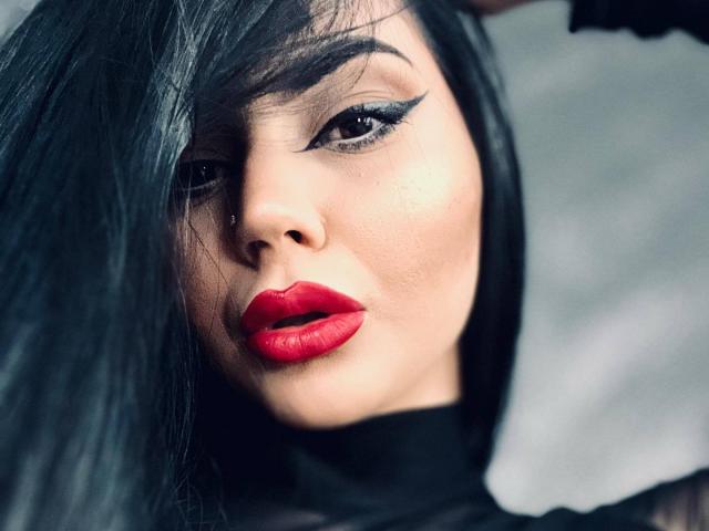 MistressEdithFuchs a les cheveux noirs, du rouge à lèvres, et un trait d'eye-liner noir épais, avec une expression sérieuse, et elle porte un haut noir. Son visage est légèrement incliné, avec les cheveux devant..