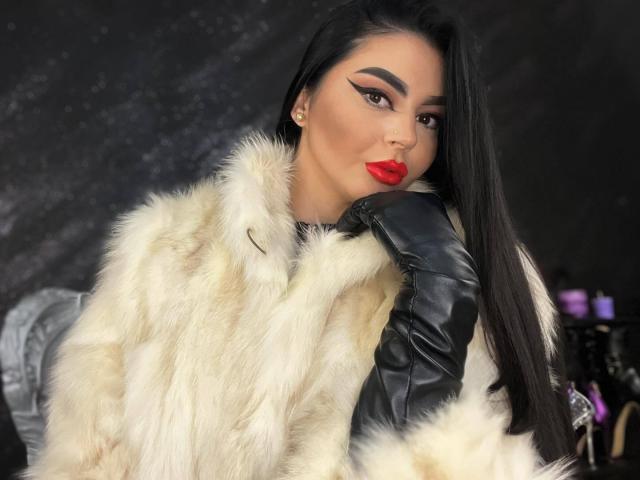 Mistress Edith Fuchs, con lunghi capelli neri, indossa un cappotto bianco di pelliccia e guanti di pelle nera, rossetto rosso e un trucco audace, appare sexy e sicura di sé. Il seno è coperto dal cappotto..
