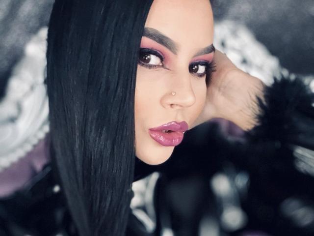 MistressEdithFuchs mit schwarzen Haaren, rosa Lidschatten und glänzenden rosa Lippen, mit einem Nasenring, die Hand am Kopf, trägt eine pelzige schwarze Jacke..