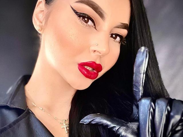 MistressEdithFuchs, avec du rouge à lèvres, des gants en cuir noir et de longs cheveux noirs, exhibe son anneau de nez et lève un doigt ganté. Elle regarde la caméra de manière séduisante..