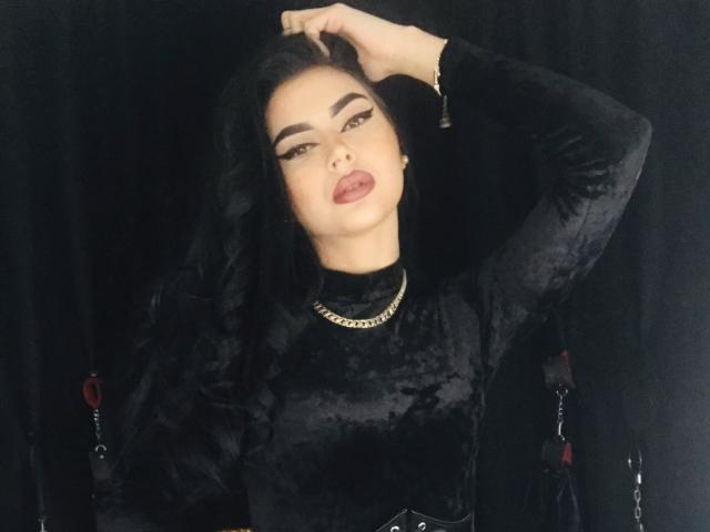 MistressEdithFuchs, met zwart, golvend haar, draagt een zwarte fluwelen top, een gouden ketting en rode lippenstift. Ze heeft haar hand in haar haar, ziet er verleidelijk uit en heeft haar mond lichtjes open..