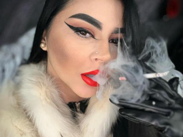 MistressEdithFuchs com um cigarro aceso, batom vermelho, delineador marcante, sobrancelhas pretas, brincos de pérolas, casaco de pele branca, luva preta, e fumaça enrolando ao redor do rosto..
