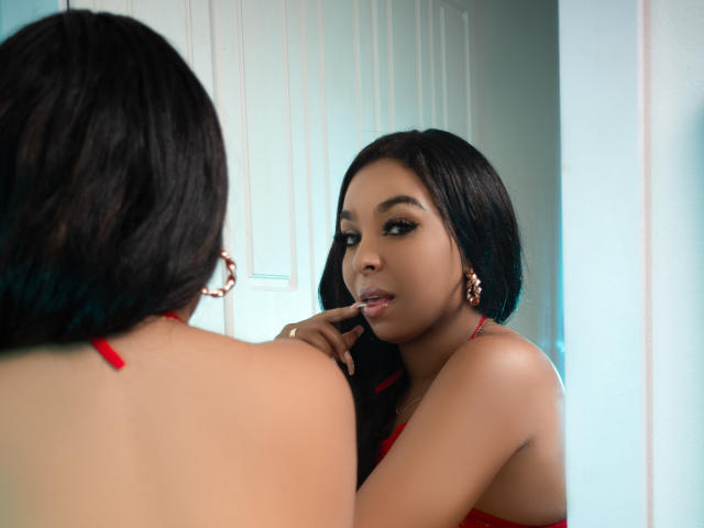 EileenBenzon with dark hair, red halter top, looking at herself in the mirror, finger on her lips, wearing gold earrings, reflecting in a white door.