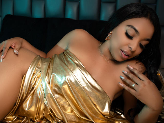 EileenBenzon is laying on a black couch, wearing a shiny gold dress, with her hand on her ass and her eyes closed, showing a relaxed, sexy expression.
