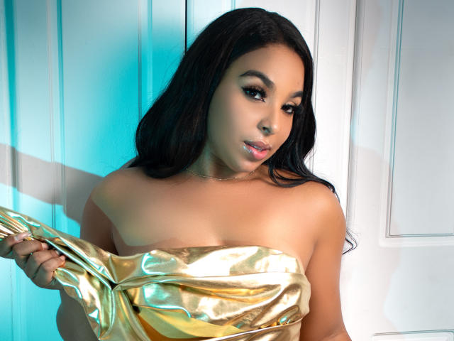 EileenBenzon with golden metallic dress, holding it over her tits, has long black hair, light brown skin, and a subtle smile. Her eyes are slightly down, looking to the side.