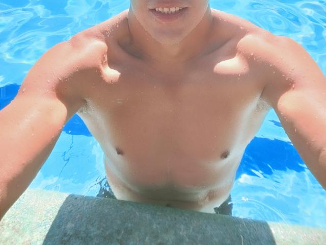 CaballeroDeLaNoches with a muscular, tan, wet, shirtless upper body, blue water, and a slight smile, standing at the pool edge, visible arms and chest, water droplets.