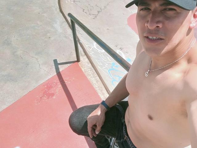 Shirtless CaballeroDeLaNoches with a medium build, wearing a black cap and necklace, sitting on a skate ramp, black pants, blue wristband, and a slight smile.