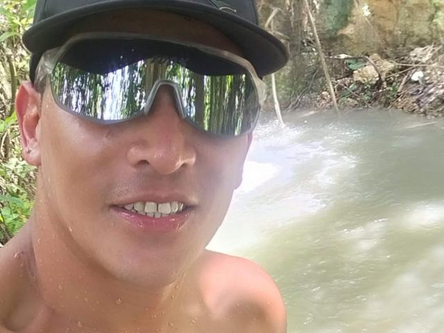 CaballeroDeLaNoches with a tan, sweaty face, white teeth, and a black cap, wearing reflective sunglasses, shirtless, in a muddy river. Water droplets on his skin.