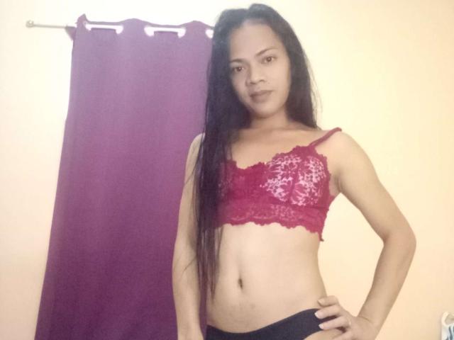 NewAsianCock with long black hair, wearing a red lace bra, showing small tits, and black pants, standing with hands on hips, looking at the camera.