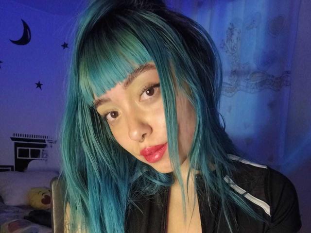 GigiVolkova with teal blue hair, nose ring, and red lipstick, looking down with a slight tilt, wearing a black shirt with white stripes, in a blue-lit room.