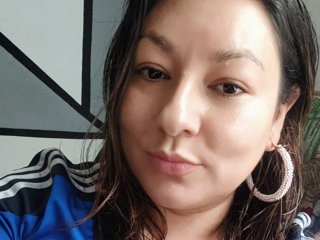 SakotoDika's face, with wet brown hair, pink hoop earrings, and a slight pout, wearing a blue and black sports jacket. Her brown eyes are slightly upturned.