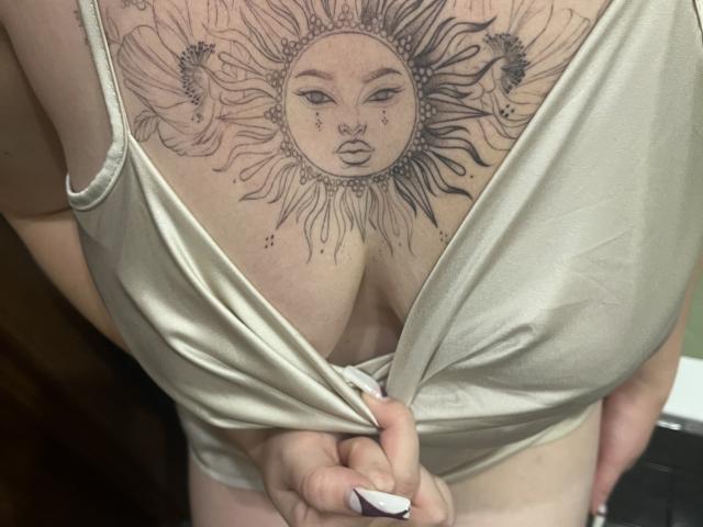DiLaria's tan tank top is pulled down, showing her large tits with a sun tattoo above her cleavage, featuring a face and rays. Her hands grip the tank top.
