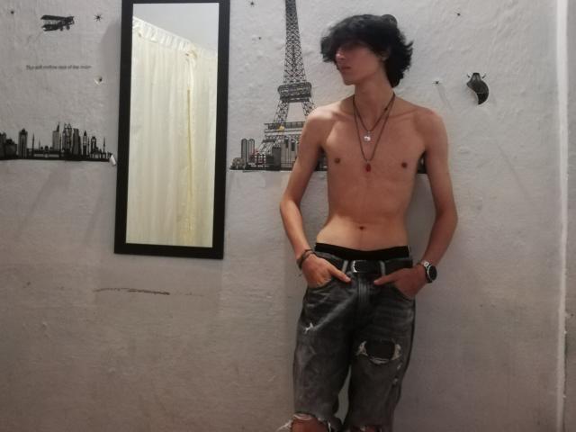 Asmodues, zonder shirt, met een slank postuur, zwart golvend haar, gekleed in versleten jeans, een ketting en een horloge, staand tegen een witte muur, met een spiegel aan de linkerkant, en een muurschildering van een stad en de Eiffeltoren..