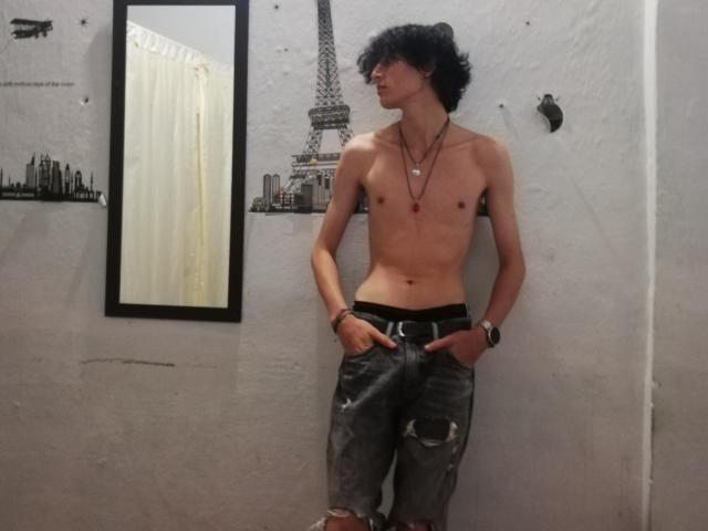 Asmodues sans chemise, avec des cheveux noirs bouclés, une silhouette mince, et des jeans déchirés, les mains dans les poches, portant des colliers et une montre, appuyé contre un mur blanc..