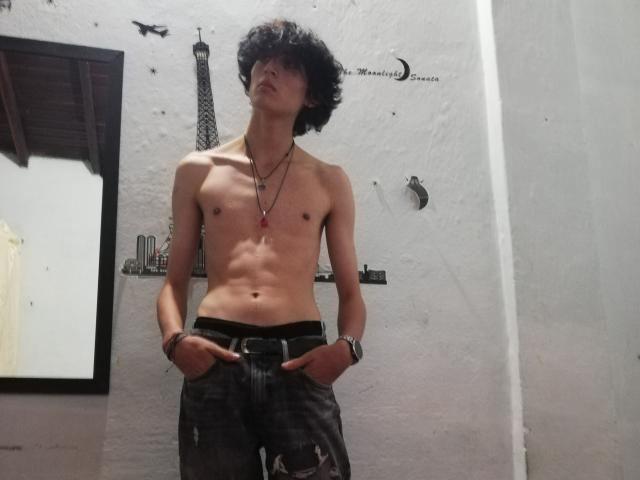 Shirtless Asmodues with curly dark hair, skinny, toned, flat ass, wearing black jeans, hands in pockets, multiple necklaces, wristbands, standing against white wall, looking up.
