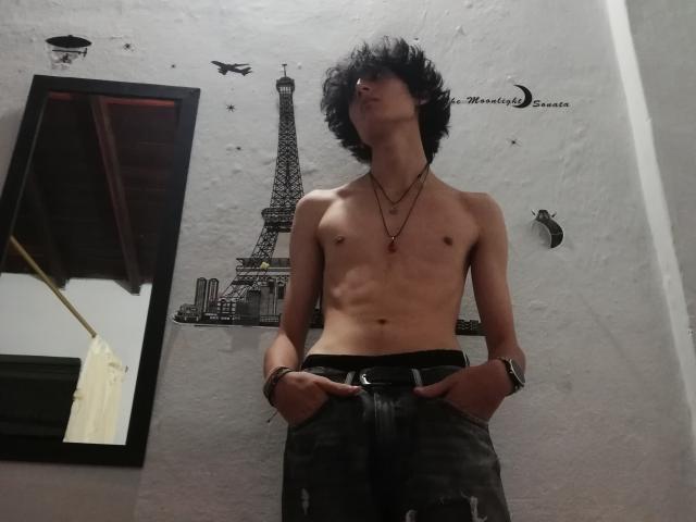 Asmodues torse nu, avec des cheveux noirs en désordre, mince avec des abdominaux visibles, portant un pantalon noir, les mains dans les poches, un collier et une montre, debout devant une œuvre d'art murale représentant la Tour Eiffel..