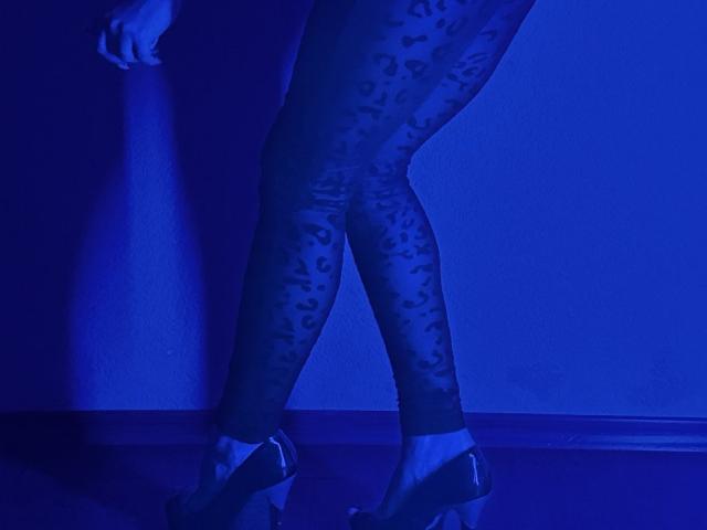 BalletSwan's legs in sheer leopard-print tights, standing in dark blue high heels, with one hand against a wall, under strong blue lighting.