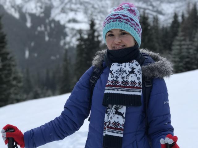 KaraCriegs with a blue jacket, red gloves, white snowflake scarf, pink and blue hat, and a black scarf, standing in snowy forest, smiling. Snow-covered mountains in background.