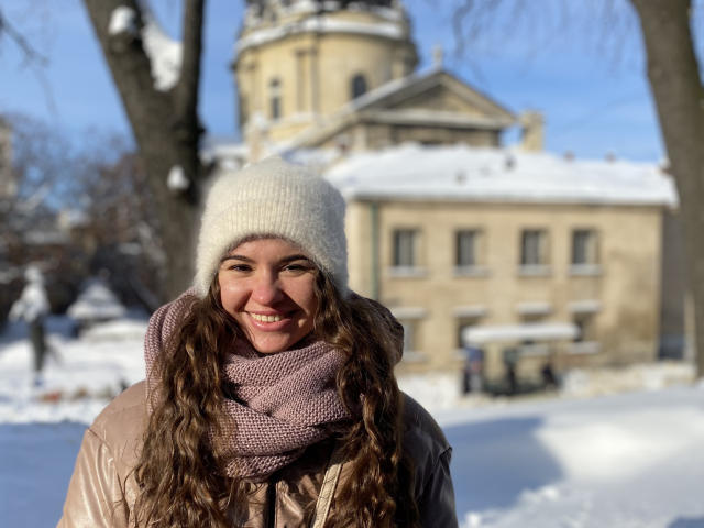 KaraCriegs met golvend bruin haar, lachend, draagt een witte gebreide muts, een roze sjaal en een beige jas, en staat in een besneeuwd landschap met een historisch gebouw op de achtergrond..