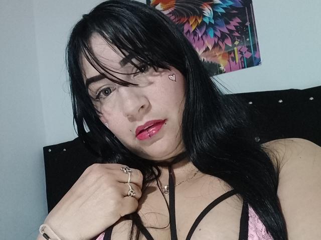 NikolSensual69 with black hair, pink lipstick, nose ring, lip ring, heart tattoo under eye, ring on finger, bracelet, pink bra, looking at camera, head slightly tilted.