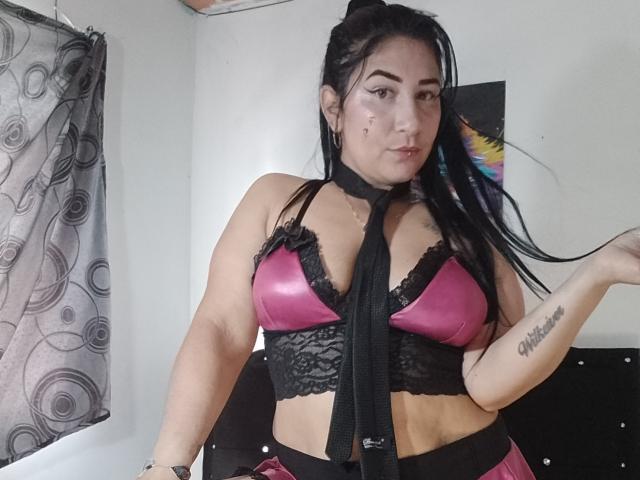 NikolSensual69 has dark hair in a ponytail, wearing a pink and black lace bra, black strap, and a tattoo on her right arm, with a confident, sexy expression.