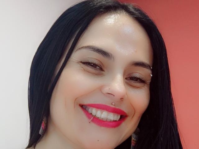 CharlotteCrofort with shiny face, black hair, pink lipstick, nose ring, lip piercing, smiling, wearing colorful earrings, and a slightly glossy forehead, against a red gradient background.