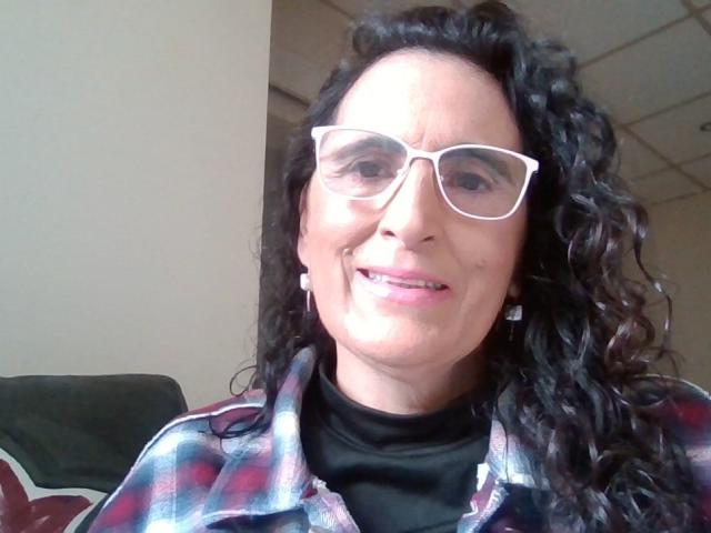 Esencia has curly black hair, white glasses, and a plaid shirt over a black turtleneck. She's smiling with pink lipstick, showing her teeth. Her ass is not visible.